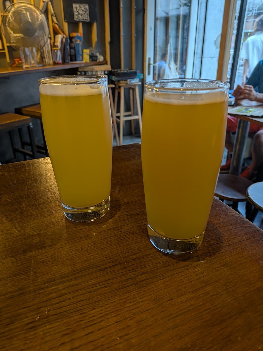 Dwie szklanki mętnego, soczystego NEIPA na barze przy oknie, z gośćmi w tle